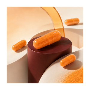 Three orange capsules and orange powder scattered on sculptural beige and dark red display blocks in a warm, minimalist studio setup.